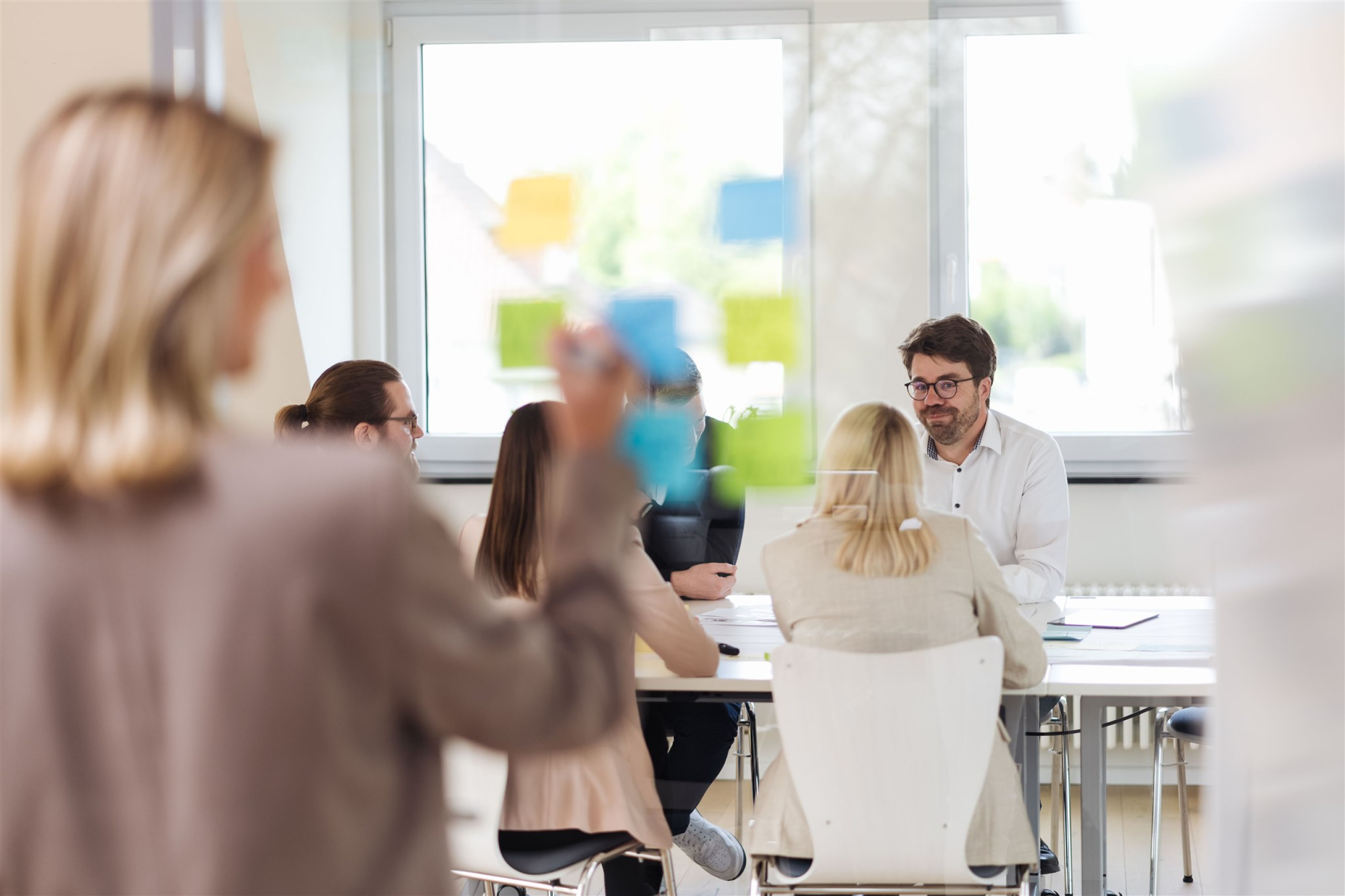 Durch eine Scheibe mit Post-Its drauf sieht man fünf Kollegen um einen Tisch herum sitzen