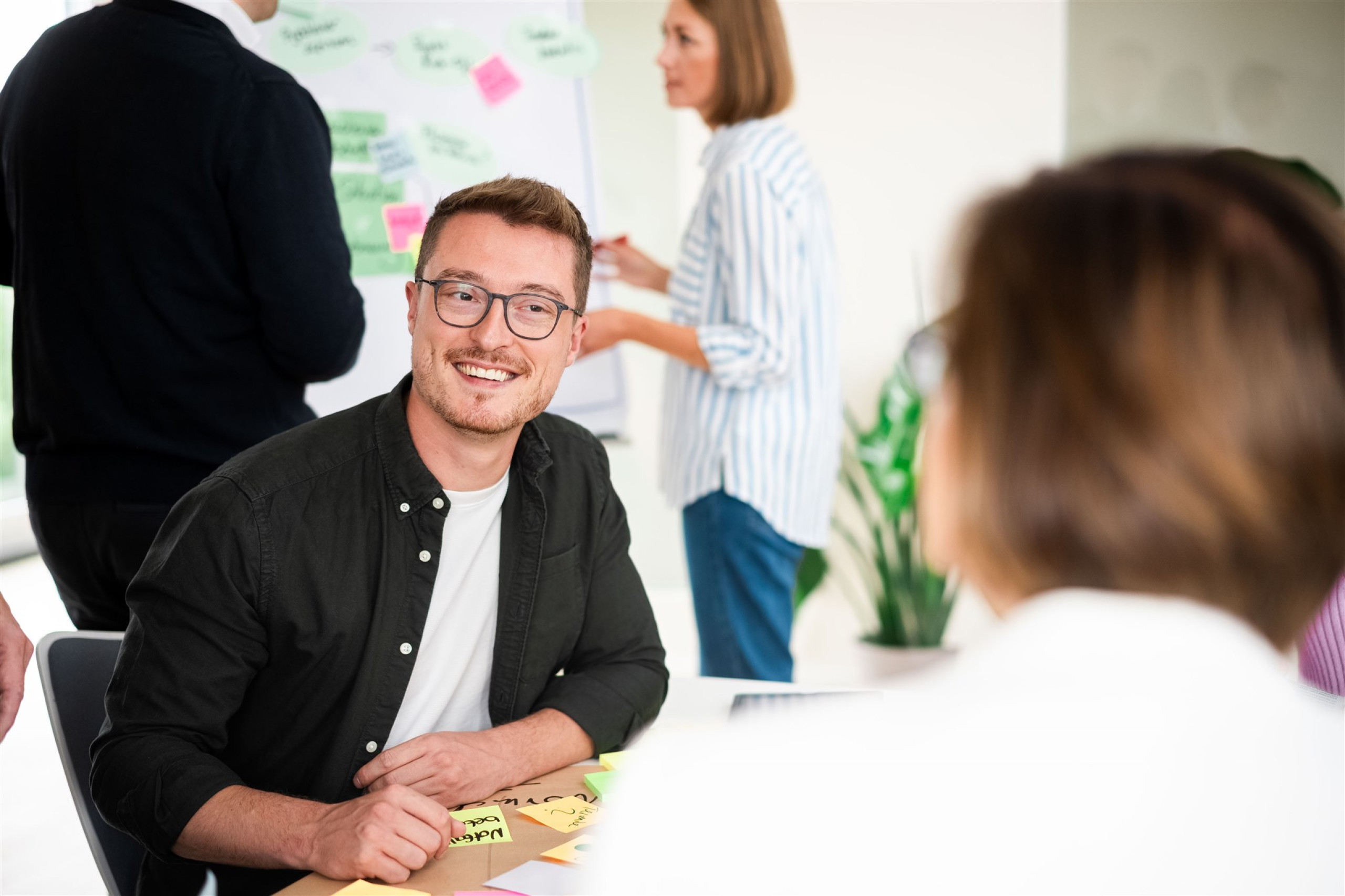 Ein Kollege lächelt eine andere Kollegin, die man nur von hinten sieht an. Es ist nur ein Ausschnitt von Kopf bis Schultern zu sehen.