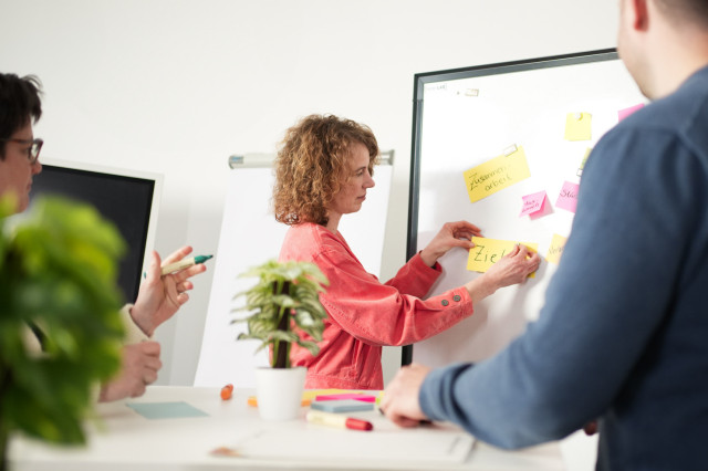 Dame klebt in einem Workshop bunte Metakarte mit der Aufschrift "Ziel"auf ein Whiteboard