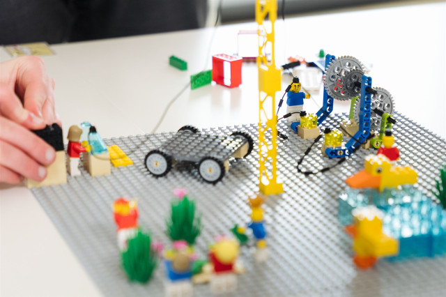 LEGO Bausteine liegen auf einem Tisch und werden von einer Hand zusammengebaut. man erkennt ein Lego-Auto und Lego-Menschen