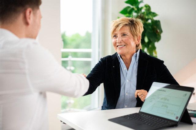 Beratungsgespräch zwischen einer Kundin und einem Berater. Beide sitzen sich am Tisch gegenünber. Die Kunden lächelt und reicht dem Berater die Hand.
