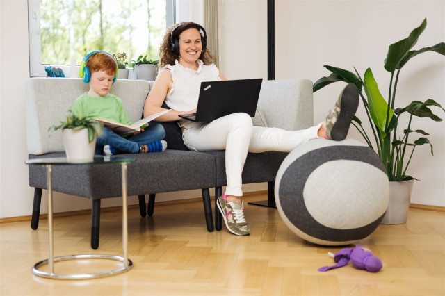 Coworking im Wonzimmer: Ein Junge und sein Vater sitzen auf dem Sofa uns arbeiten konzentriert. Der Vater an seinem Laptop und  ser Sohn  mit Kopfhörern liest ein Buch,. beide lächeln.