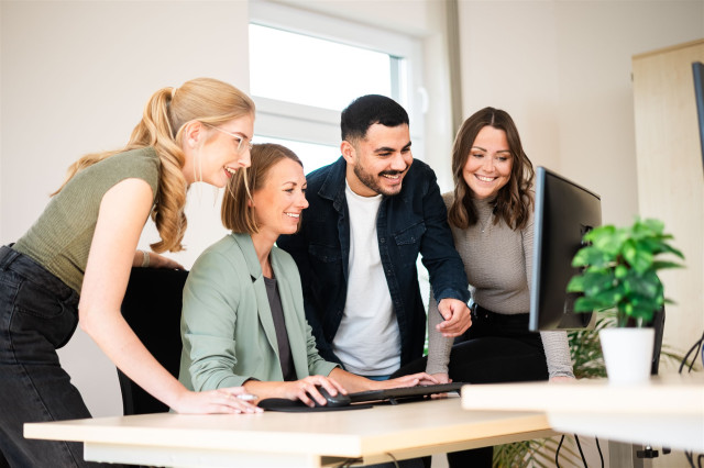 vier Mitarbeitende stehen gemeinsam vor einem Schreibtisch, sie beugen sich zueinander runter und schauen lachend auf den laptop.
