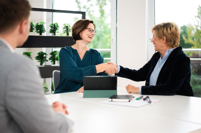 zwei Personen schütteln sich die Hand. Eine dritte steht daneben.