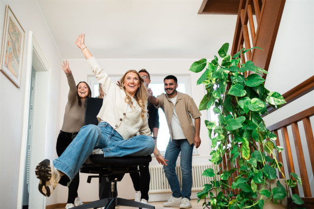 eine Kollegin sitzt auf einem Bürostuhl und wird von den anderen Kollegen angeschubst